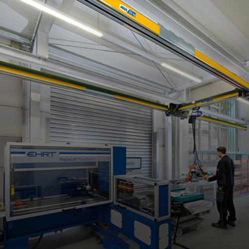 Einträgerkran EHB-X bewegt eine Last in einer Industriehalle, während ein Mitarbeiter im Hintergrund an einer Maschine arbeitet.